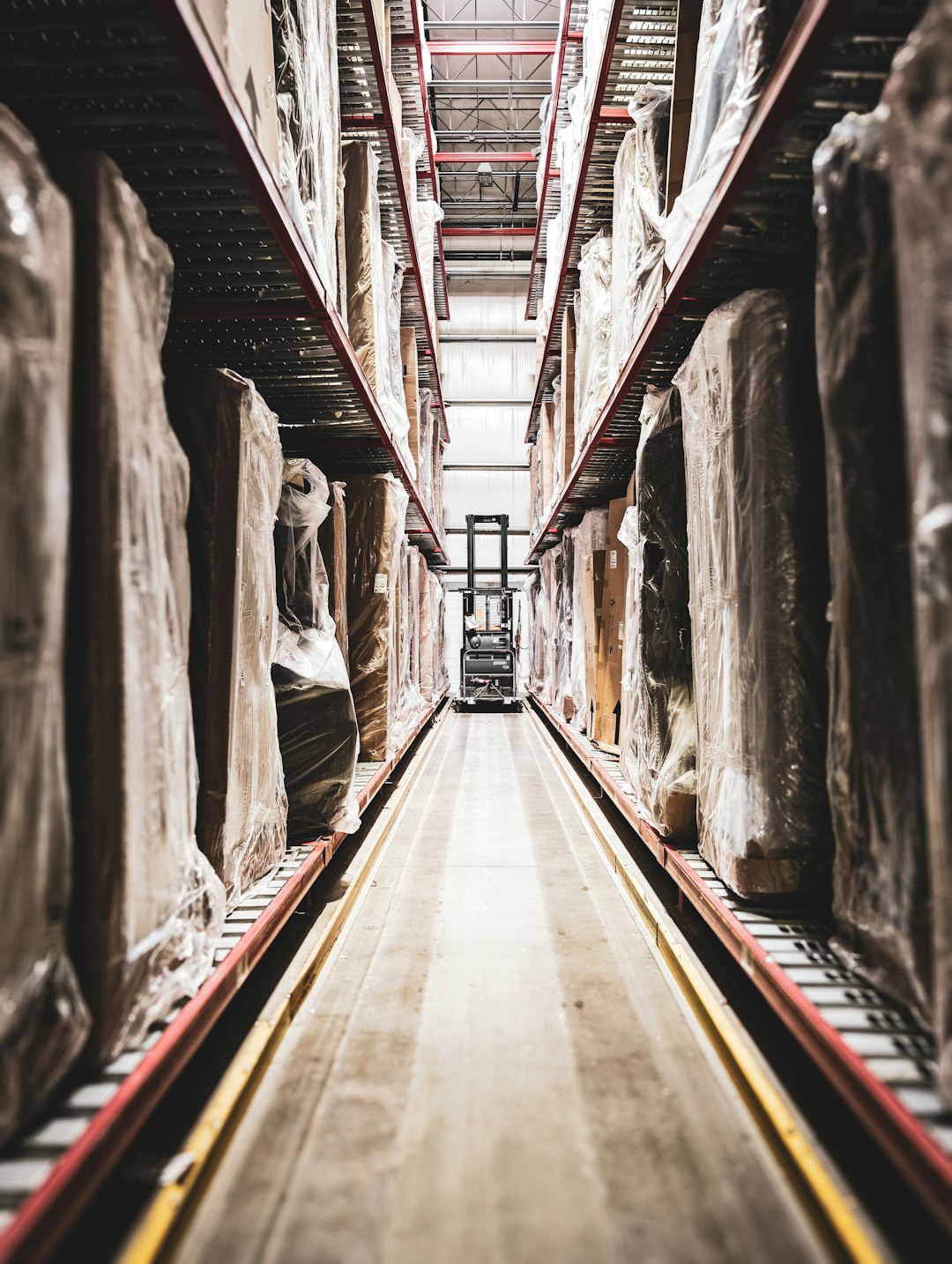 A lone forklift navigates a narrow aisle in a cavernous warehouse, dwarfed by towering shelves brimming with shrink-wrapped inventory. The scene captures the scale and precision of modern logistics, where every item has its designated place and every movement is orchestrated for maximum efficiency.