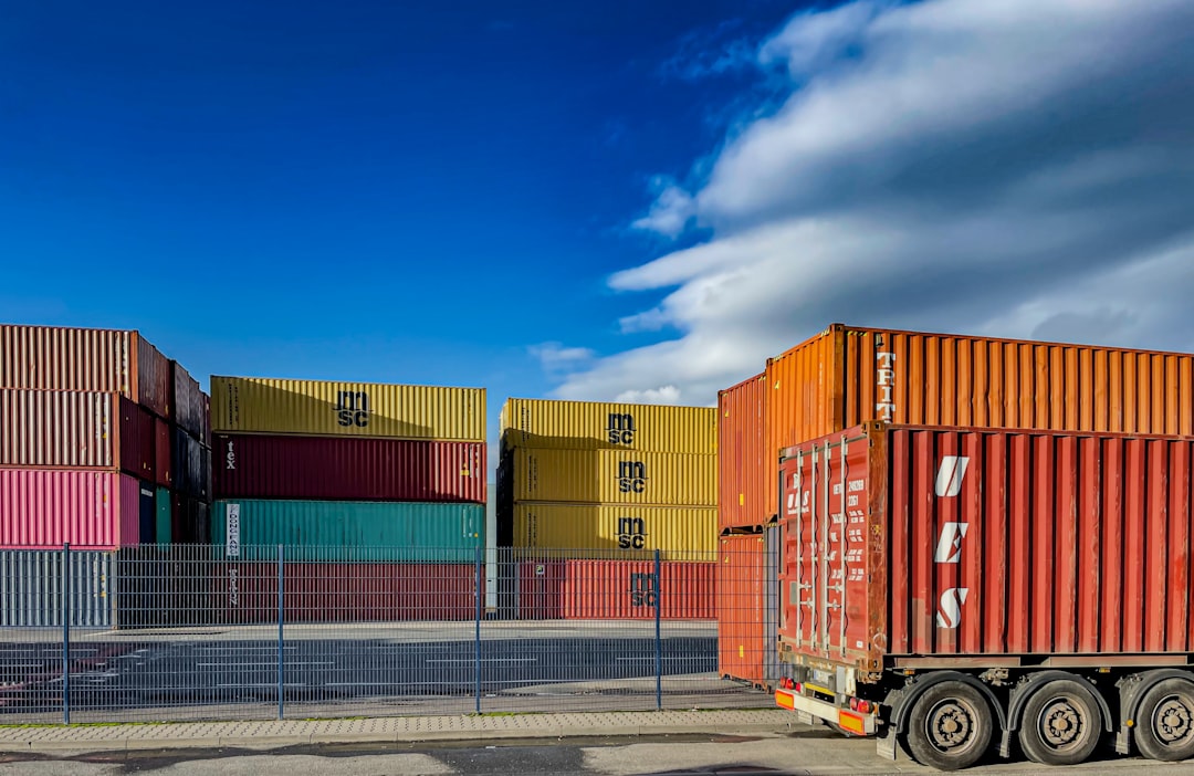 Stacked colorful shipping containers in a freight terminal, with a clear blue sky and clouds above, and one container loaded on a truck trailer in the foreground